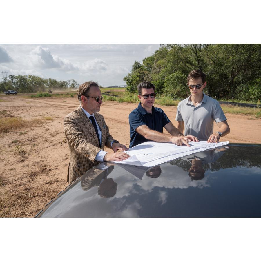 Three men in sunglasses review a large site plan spread across the hood of a car on cleared land at the Occam Foundry ca