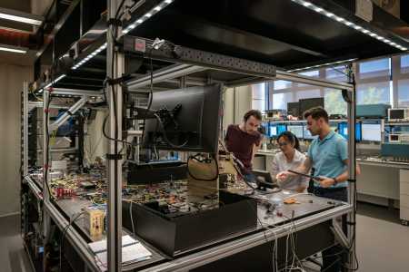 Three researchers examine a complex electronic workstation with numerous cables and components under bright lighting.