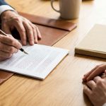 Two pairs of hands at a desk, one signing a document with a fountain pen